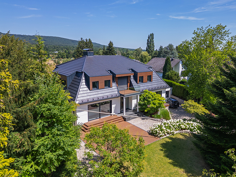 Aerial view of a modern house with a dark tiled roof, wooden accents, and a patio surrounded by lush trees and a garden.