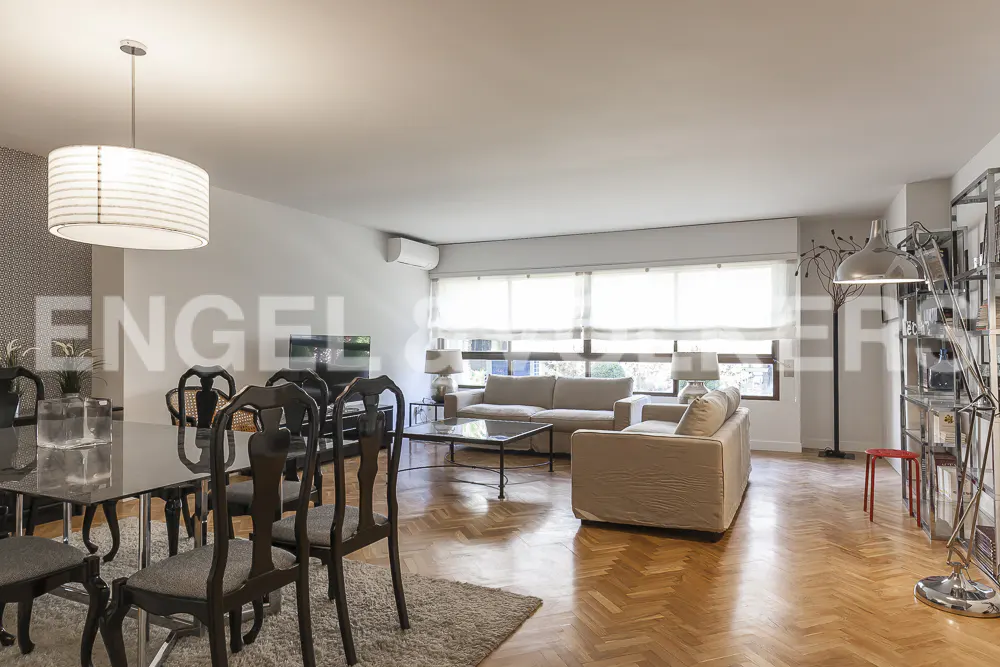 Bright, open-plan living and dining area with hardwood floors. Beige sofas, black dining table with chairs, and a large window.