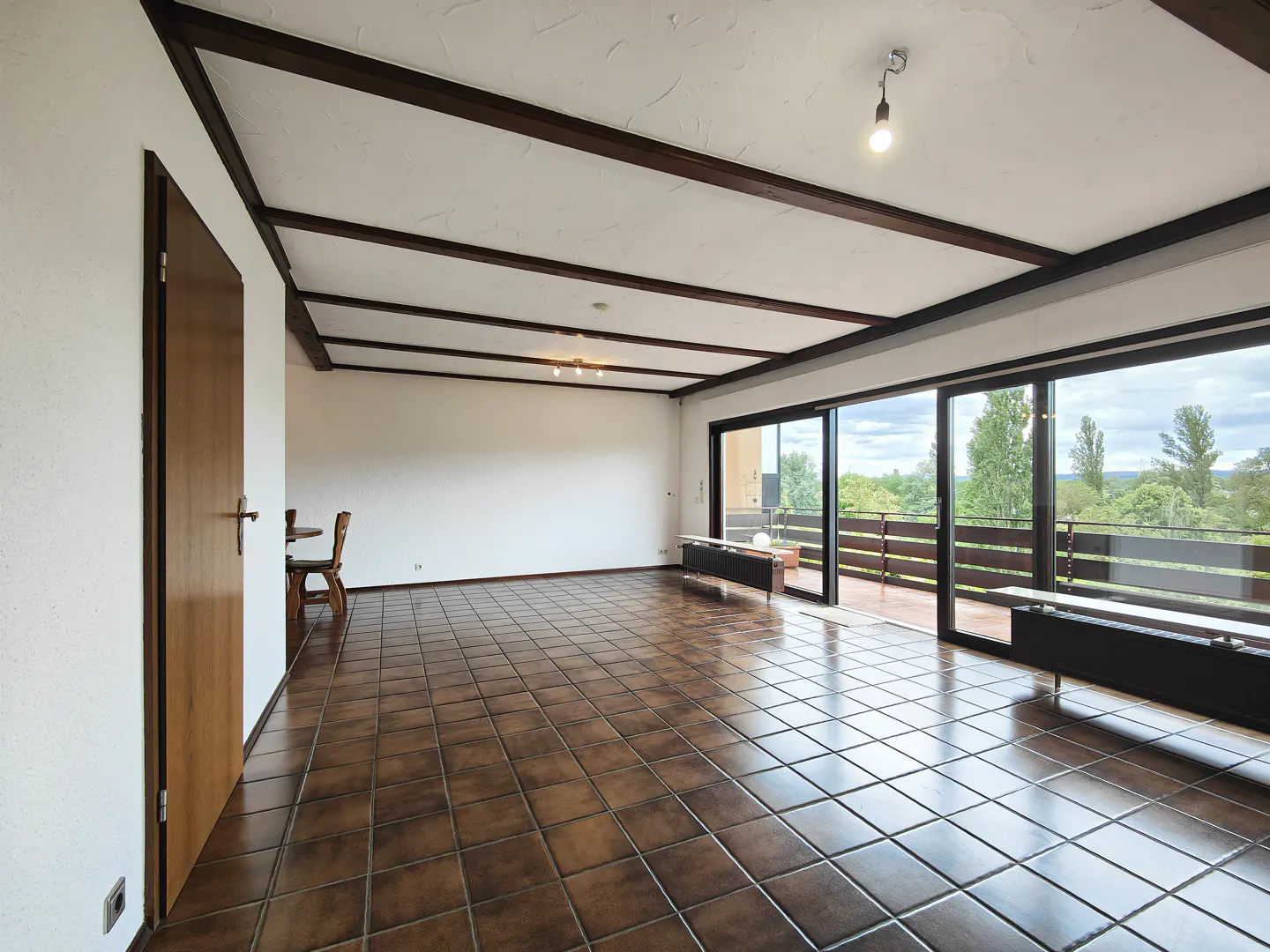 Spacious room with brown tile floor, white walls, and dark wood beams on the ceiling. Sliding glass doors lead to a balcony with a view of trees.