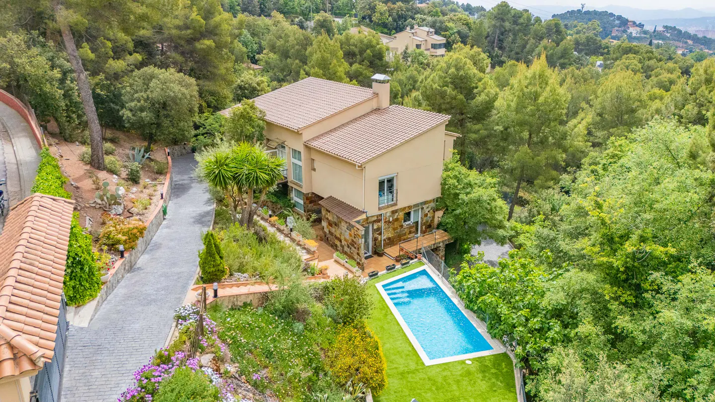 Aerial view of a beige two-story house with a brown tile roof, a blue pool, and lush green trees.