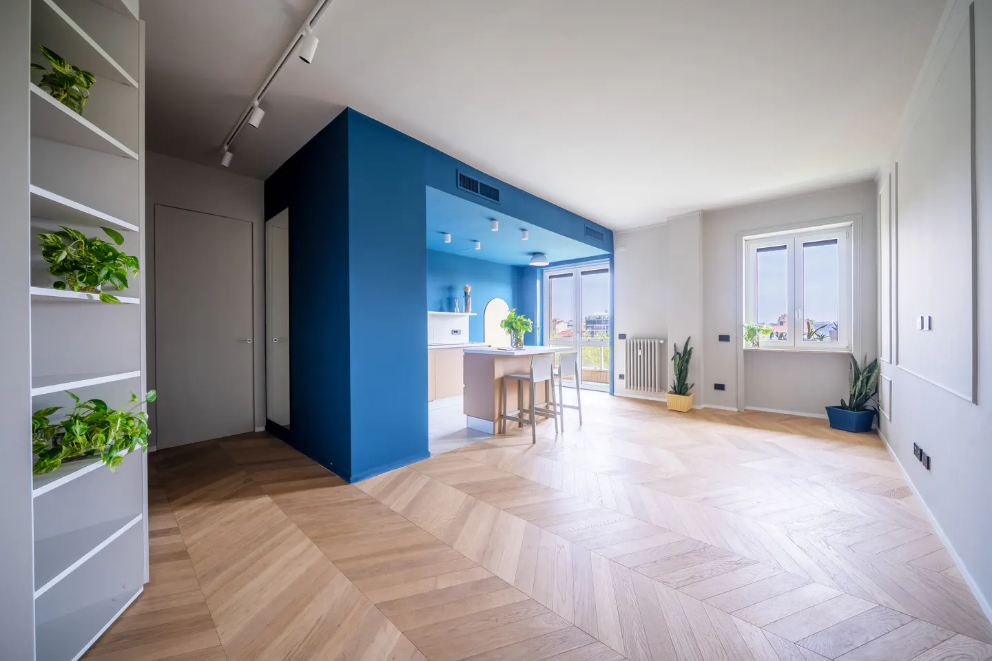 Bright, modern apartment interior with herringbone wood floors, blue accent wall, kitchen island, and large windows. Plants add a touch of green.