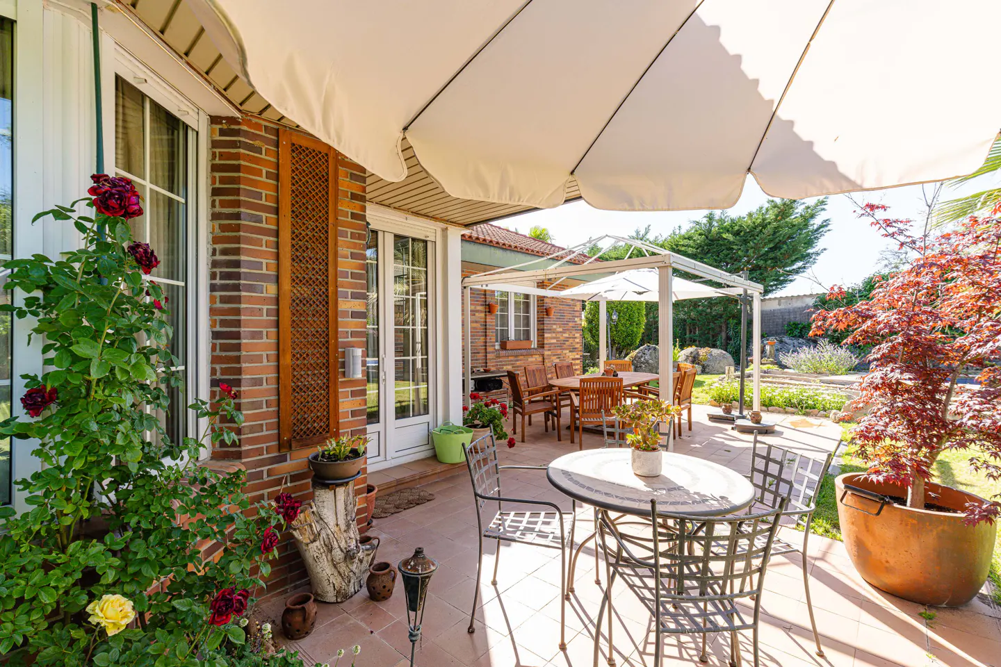 Outdoor patio with metal table and chairs, brick house, white umbrella, and garden with red maple tree.