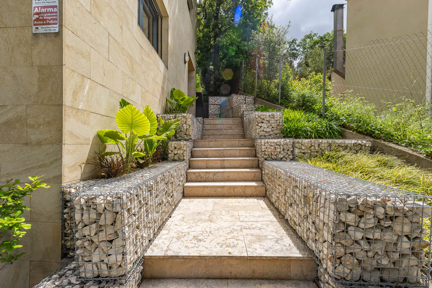Stone steps lead up to a building, flanked by wire cages filled with rocks and greenery.