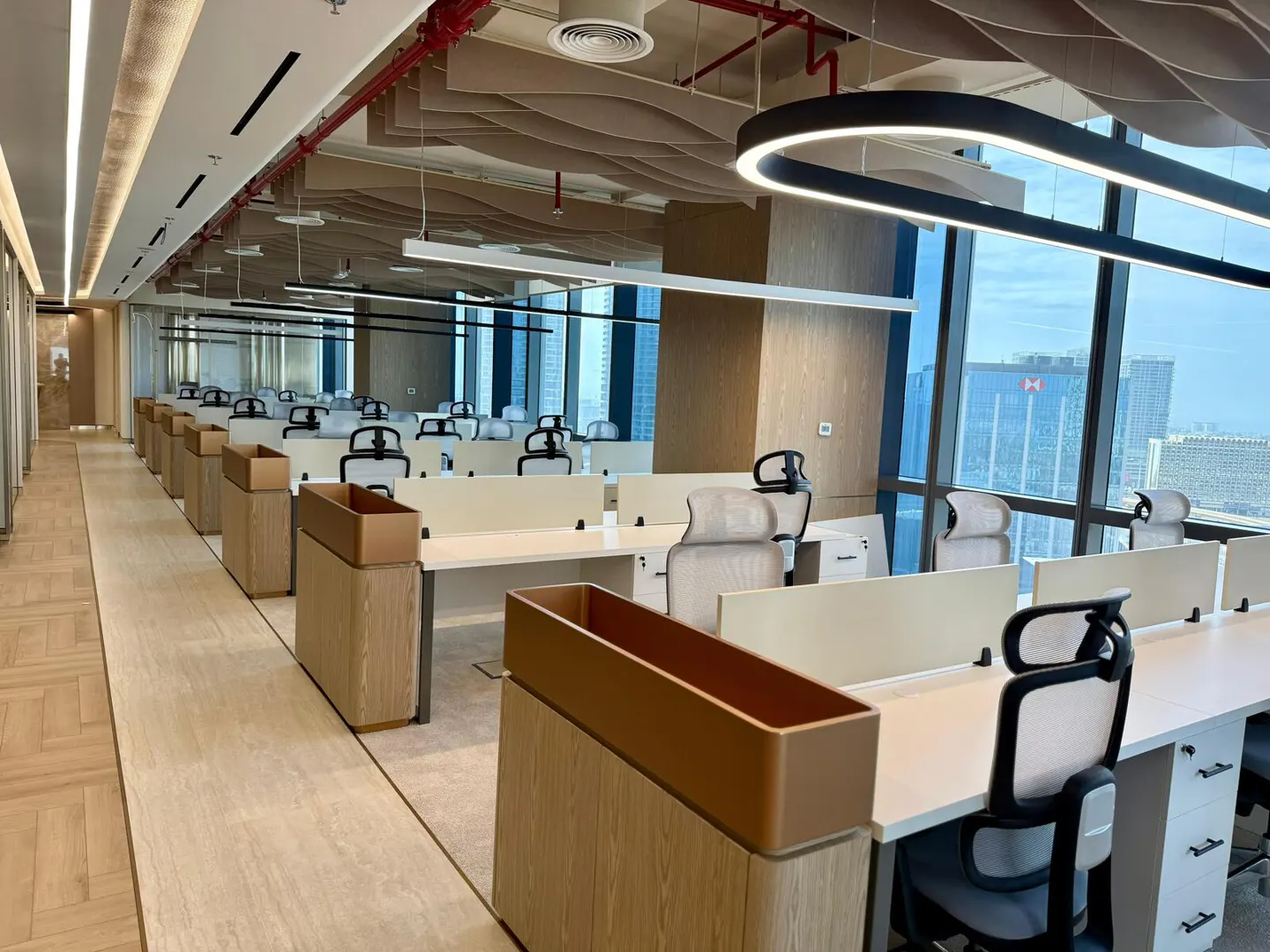 A modern office space with rows of desks and chairs, large windows, and city views.