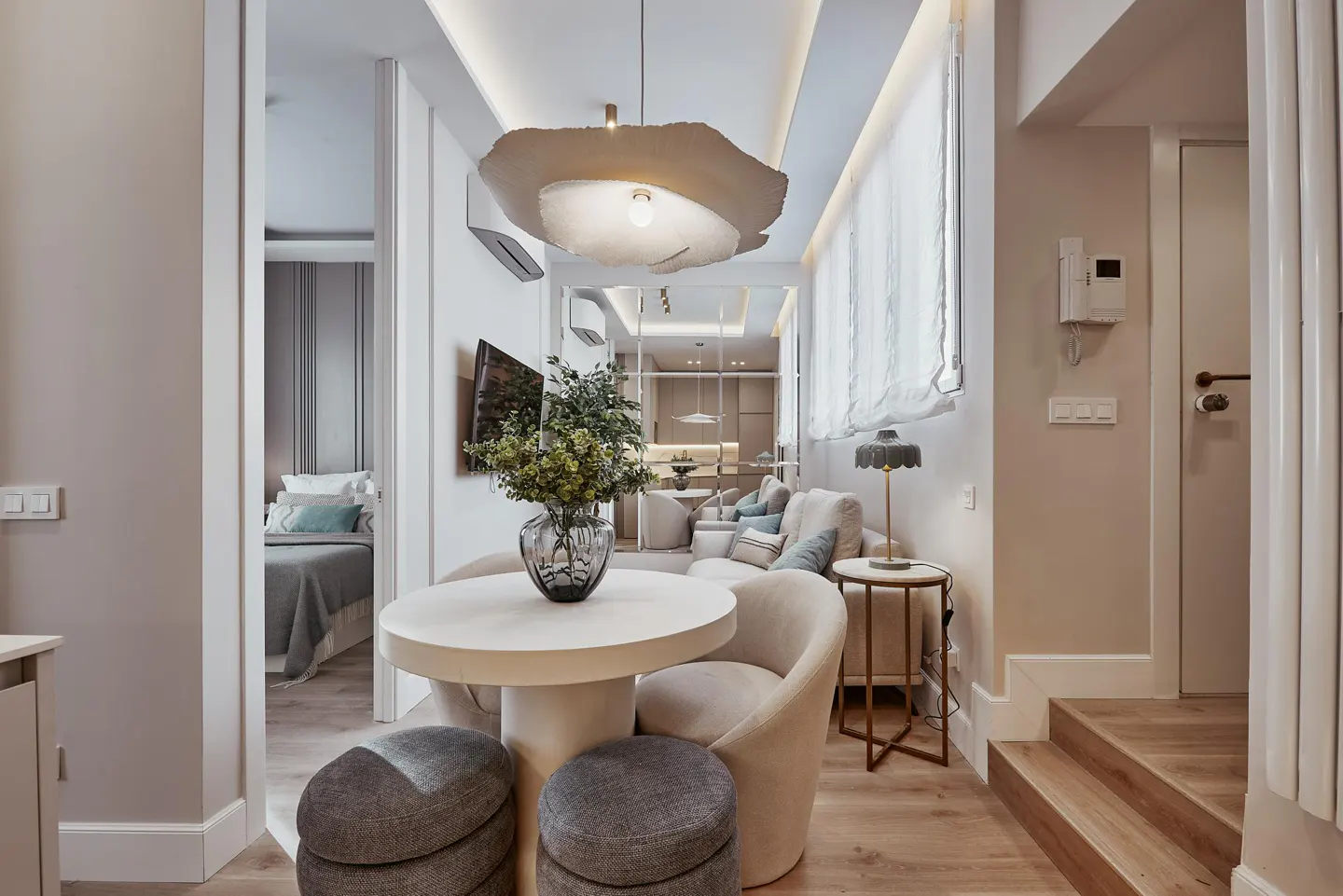 Interior view of a modern apartment with a round table, chairs, and a living area with a sofa and TV. The color palette is neutral with beige and gray tones.