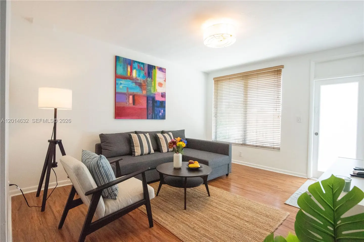 Living room with wood floors, white walls, and a gray sectional sofa. A colorful painting hangs above the sofa. A round coffee table sits on a jute rug.