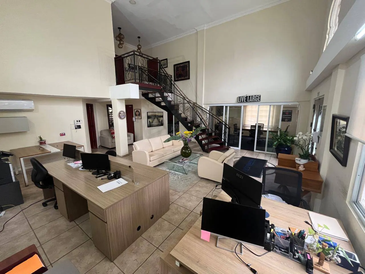 Open office space with desks, computers, and a seating area with cream sofas. A black staircase leads to a second-floor balcony. "Live Large" sign visible.