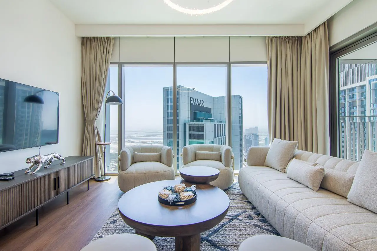 Living room with a beige sofa, two chairs, and a round coffee table. Large windows offer a city view with the Emaar building.