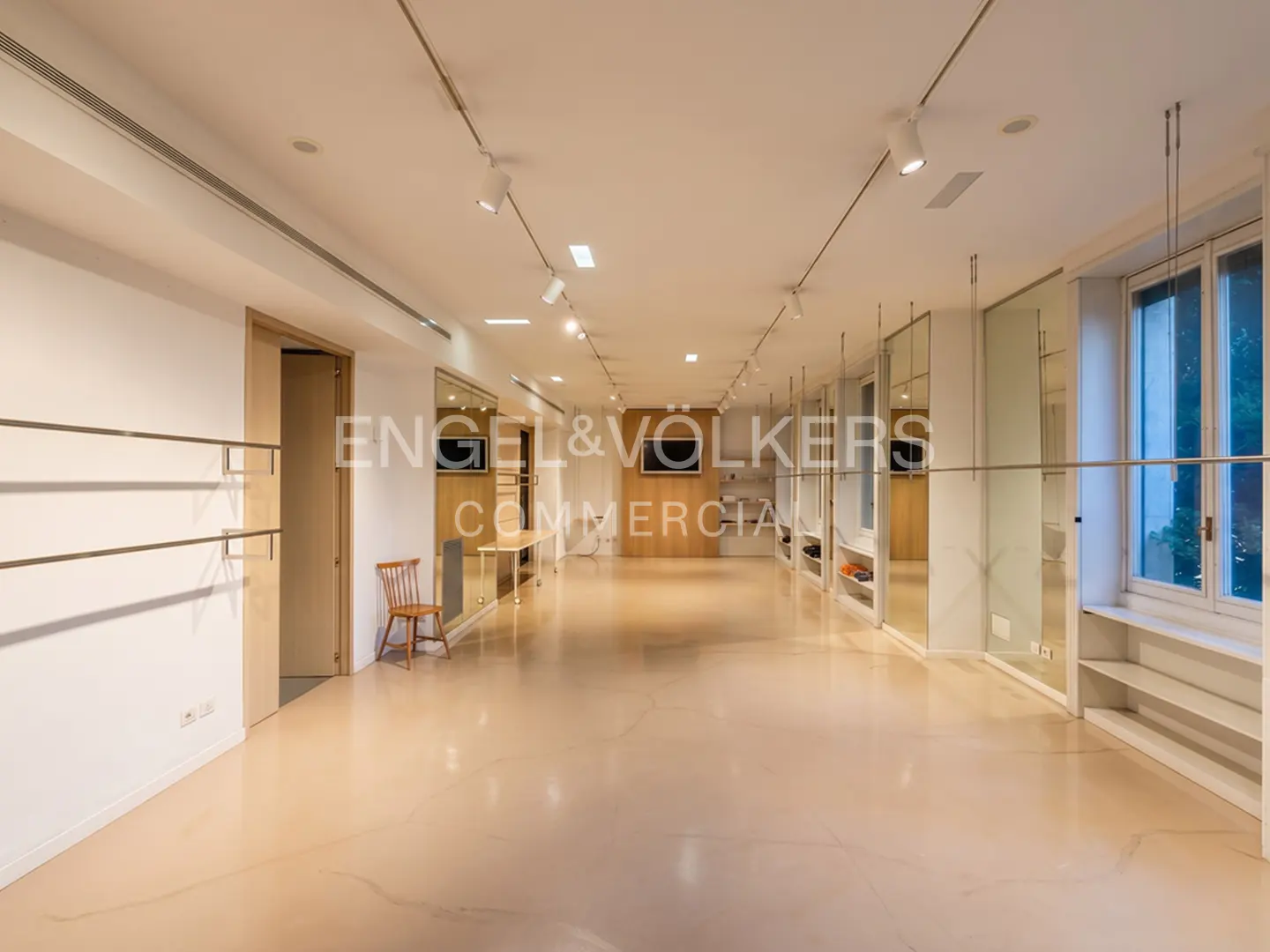 Modern, minimalist commercial space with light-colored floors, track lighting, wall shelving, and a single wooden chair.