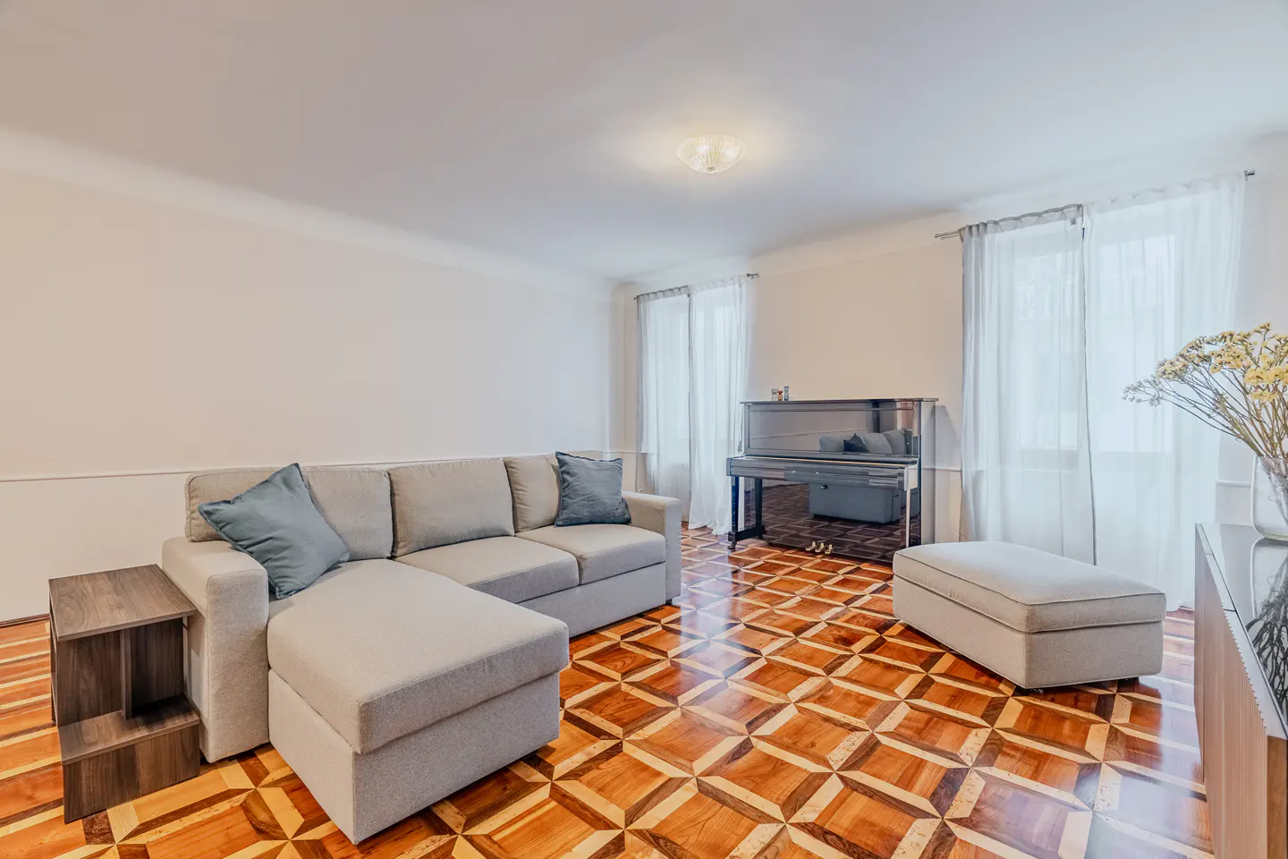 Bright living room with a light gray sectional sofa, matching ottoman, and an upright piano on patterned hardwood floors.