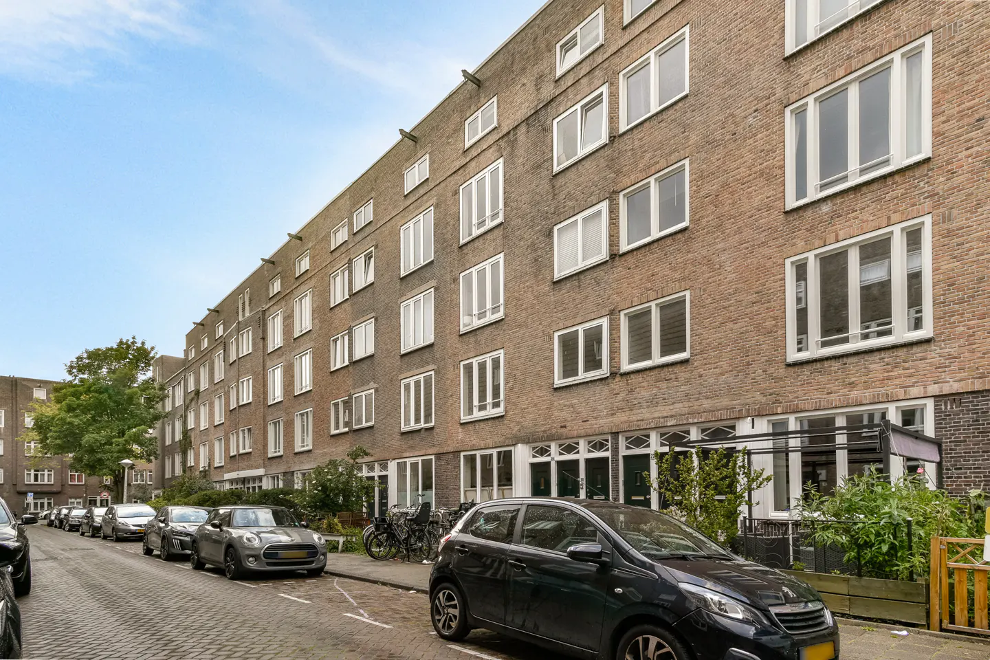 A long, brick apartment building with white-framed windows lines a street with parked cars.