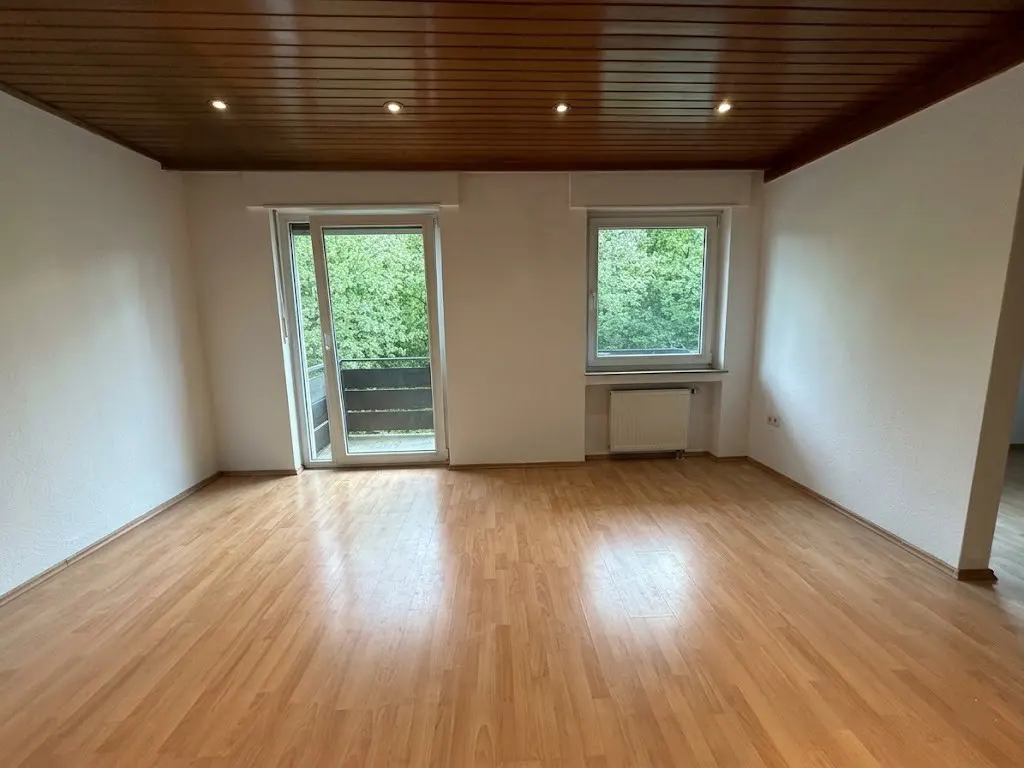 Bright, empty room with wood floors, white walls, and a wood-paneled ceiling. A sliding glass door leads to a balcony, and a window overlooks green trees.