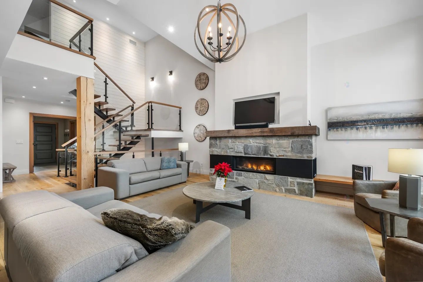 Bright living room with gray sofas, stone fireplace, and wood floors. A staircase leads to a second-floor loft.