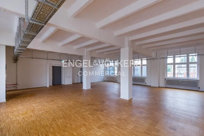 Empty industrial-style office space with white walls, exposed ceiling beams, wooden floors, and large windows.