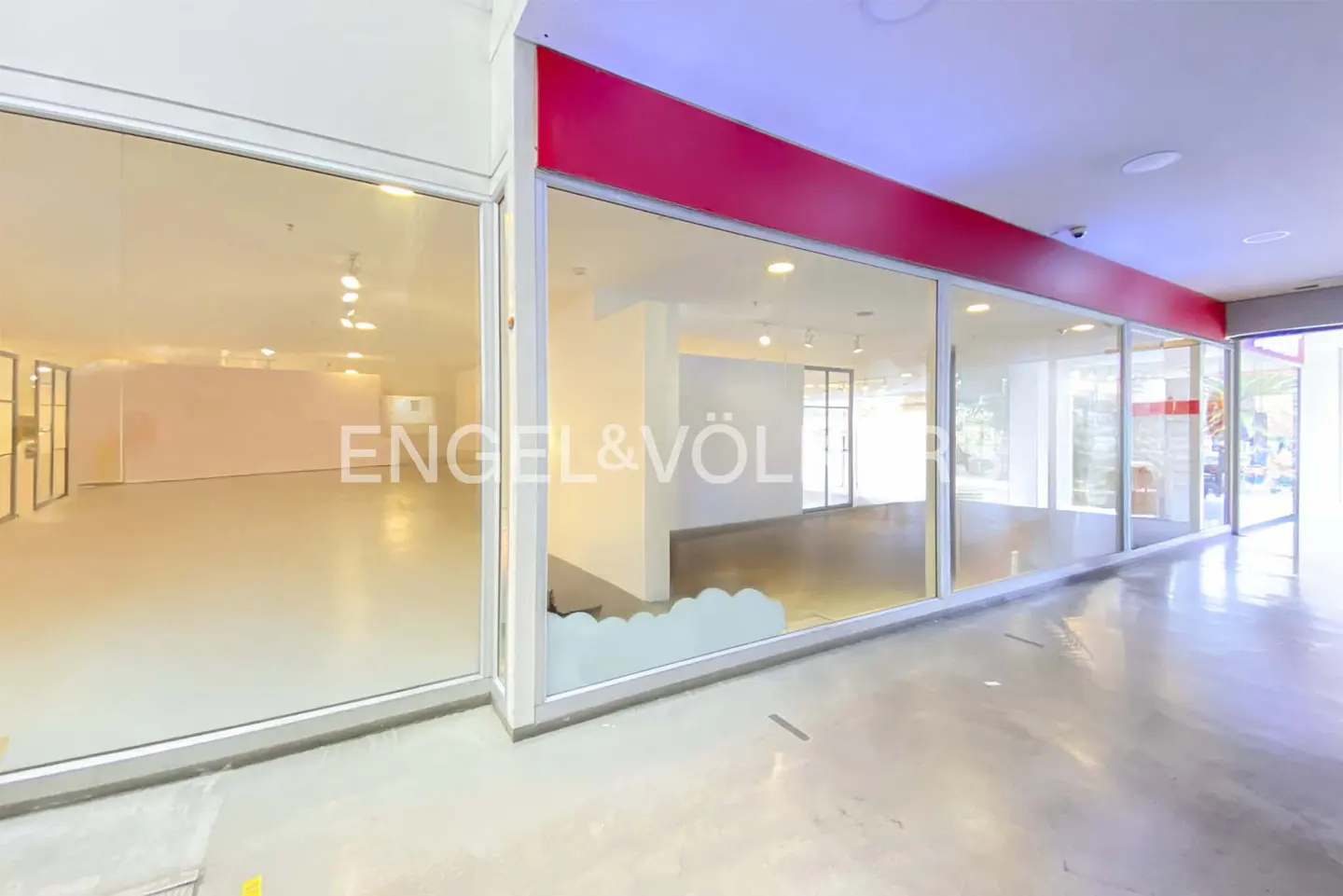 Empty retail storefront with large glass windows, white interior walls, and a prominent red sign header.