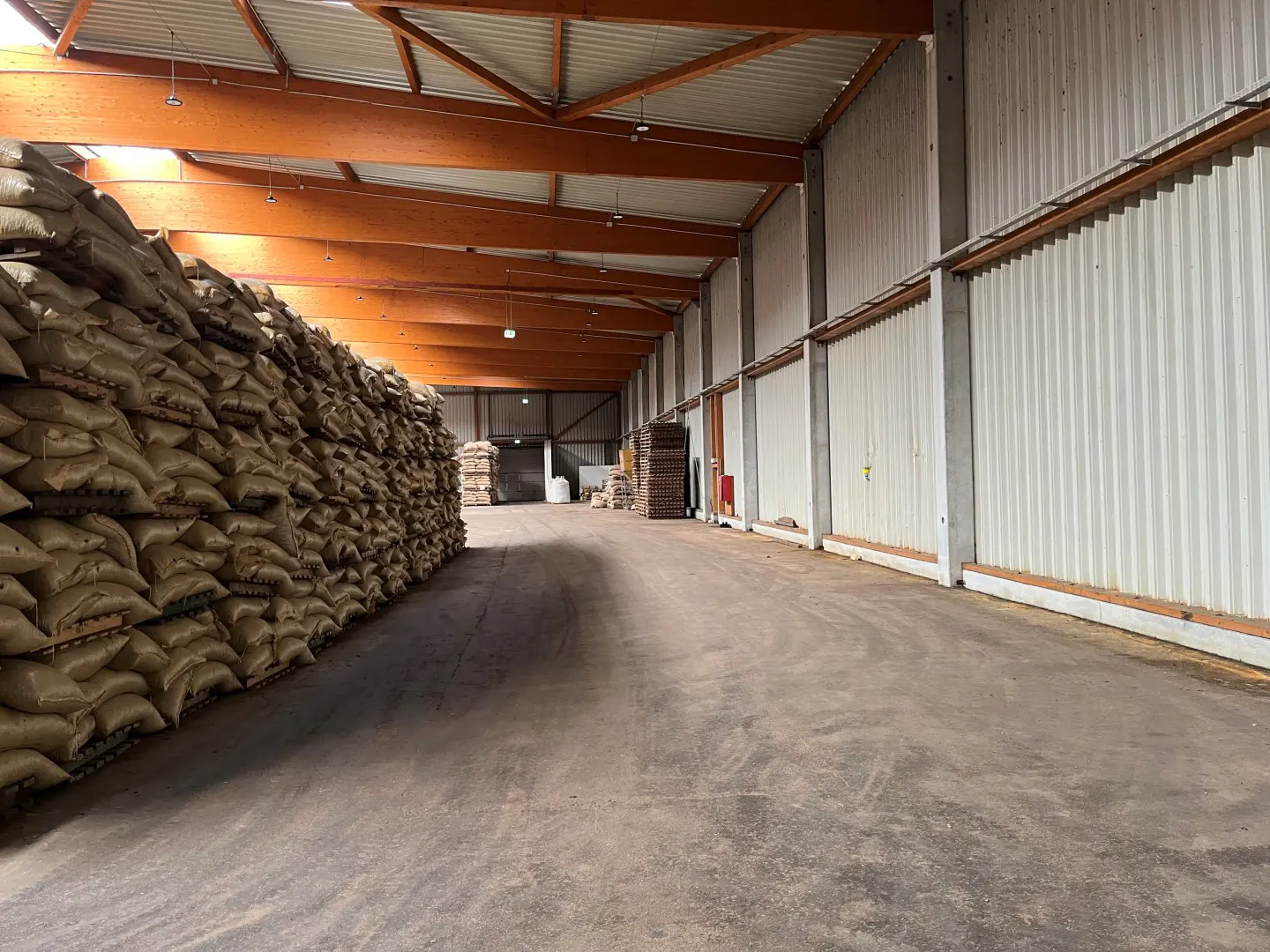 A warehouse interior with a concrete floor, metal walls, and a wooden beam ceiling. A large stack of burlap sacks is on the left.