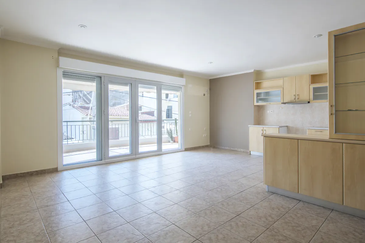 Bright, empty apartment with tile floors, beige walls, and light wood kitchen cabinets. Sliding glass doors lead to a balcony.