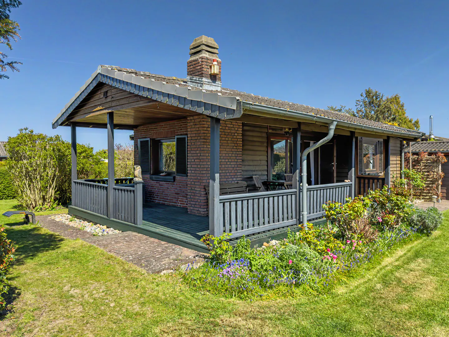 A small wooden cabin with a brick chimney and a covered porch, surrounded by a lush garden under a clear blue sky.