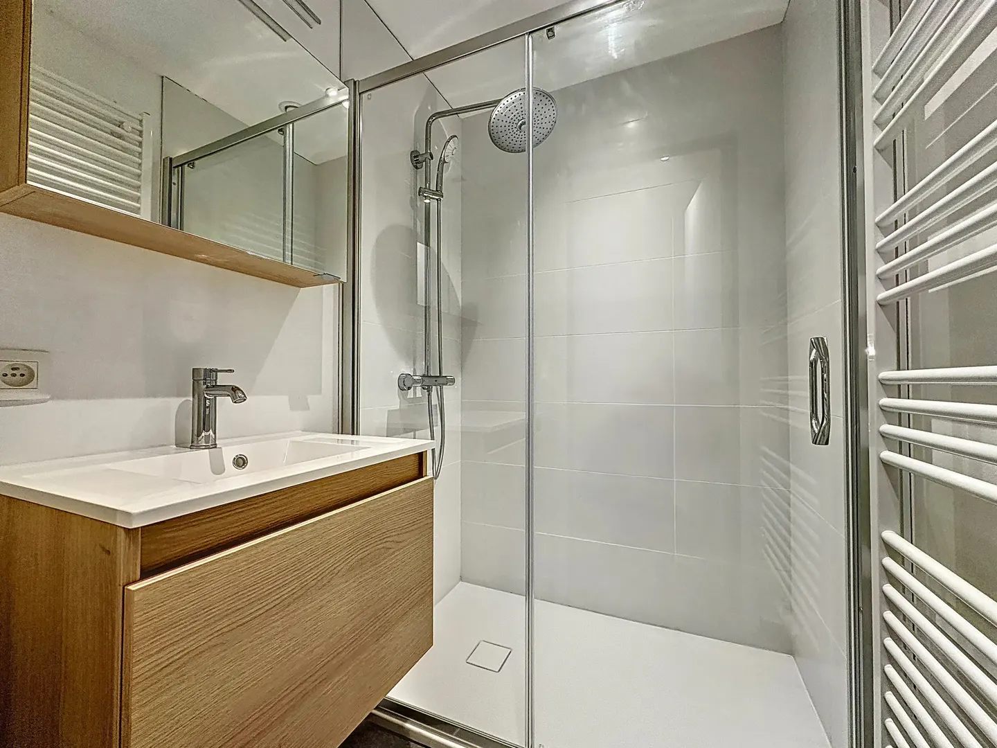 Modern bathroom with a wood vanity, white countertop, and a glass-enclosed shower with white tiles. A towel warmer is on the right.