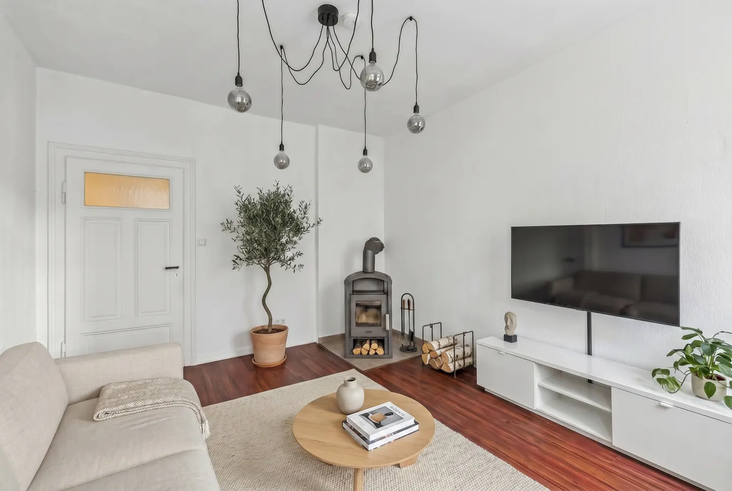 Bright living room with white walls, wood floors, and a beige sofa. A wood-burning stove sits near a potted olive tree. A modern light fixture hangs above.