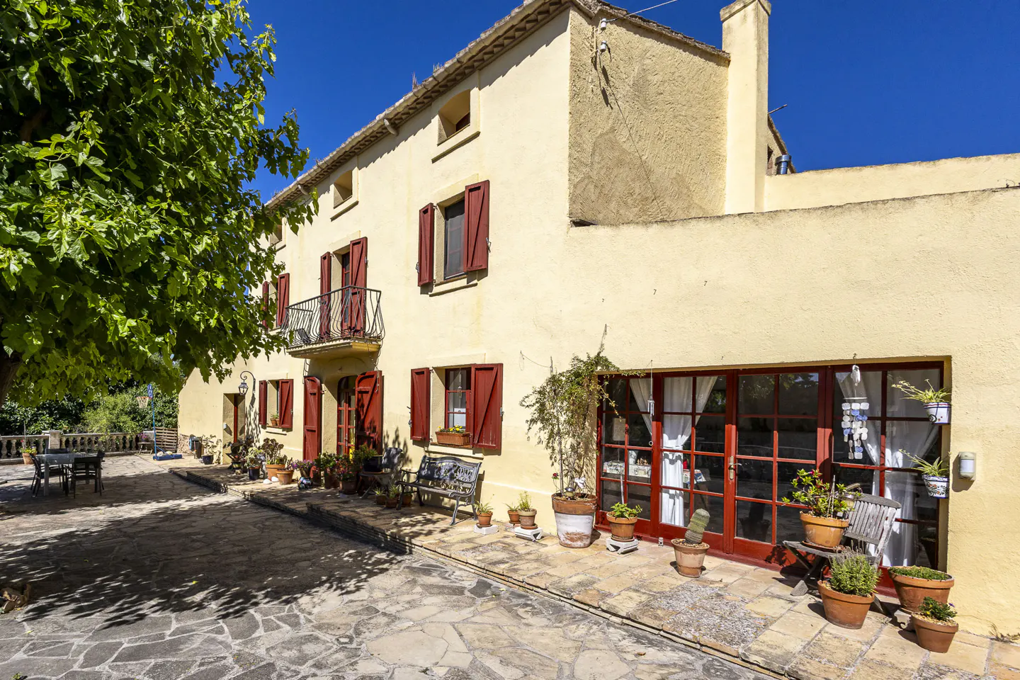 A yellow stone house with red shutters and a stone patio filled with potted plants under a bright blue sky.
