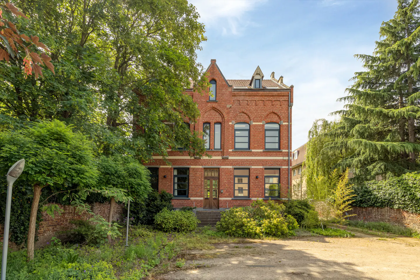 A three-story red brick house with arched windows and a wooden front door, surrounded by lush trees and a gravel driveway.