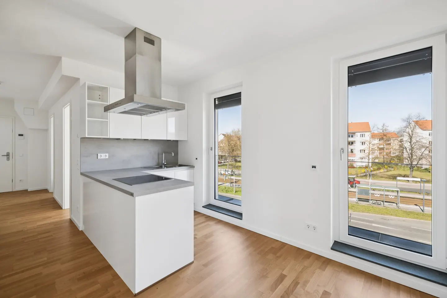 Bright, modern kitchen with white cabinets, stainless steel hood, and gray countertops. Hardwood floors and large windows with city views.