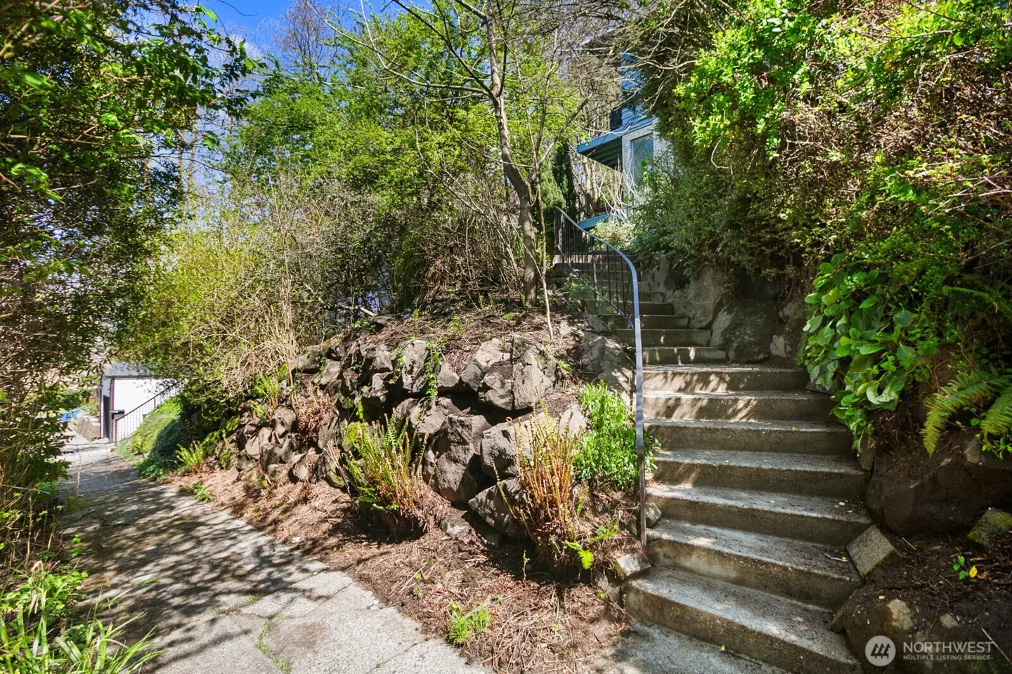 Stone steps lead up a hillside to a house, with lush greenery and a rock wall. A sidewalk branches off to the left.