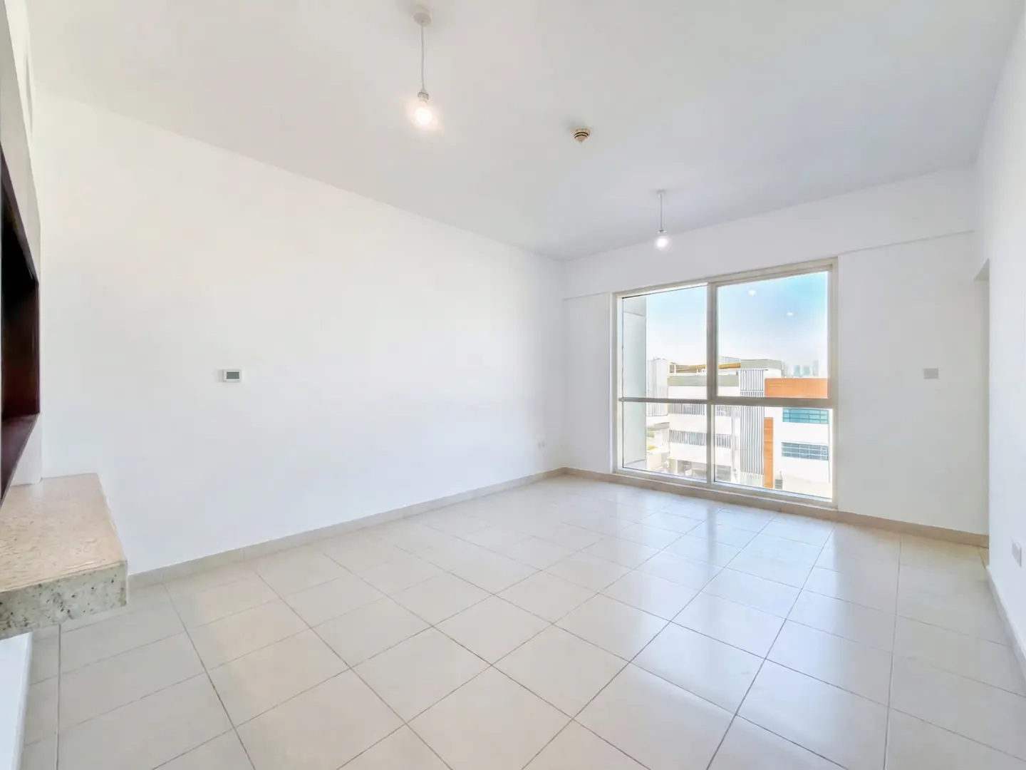 Bright, empty living room with white walls, light tile floors, two pendant lights, and a large window with city views.