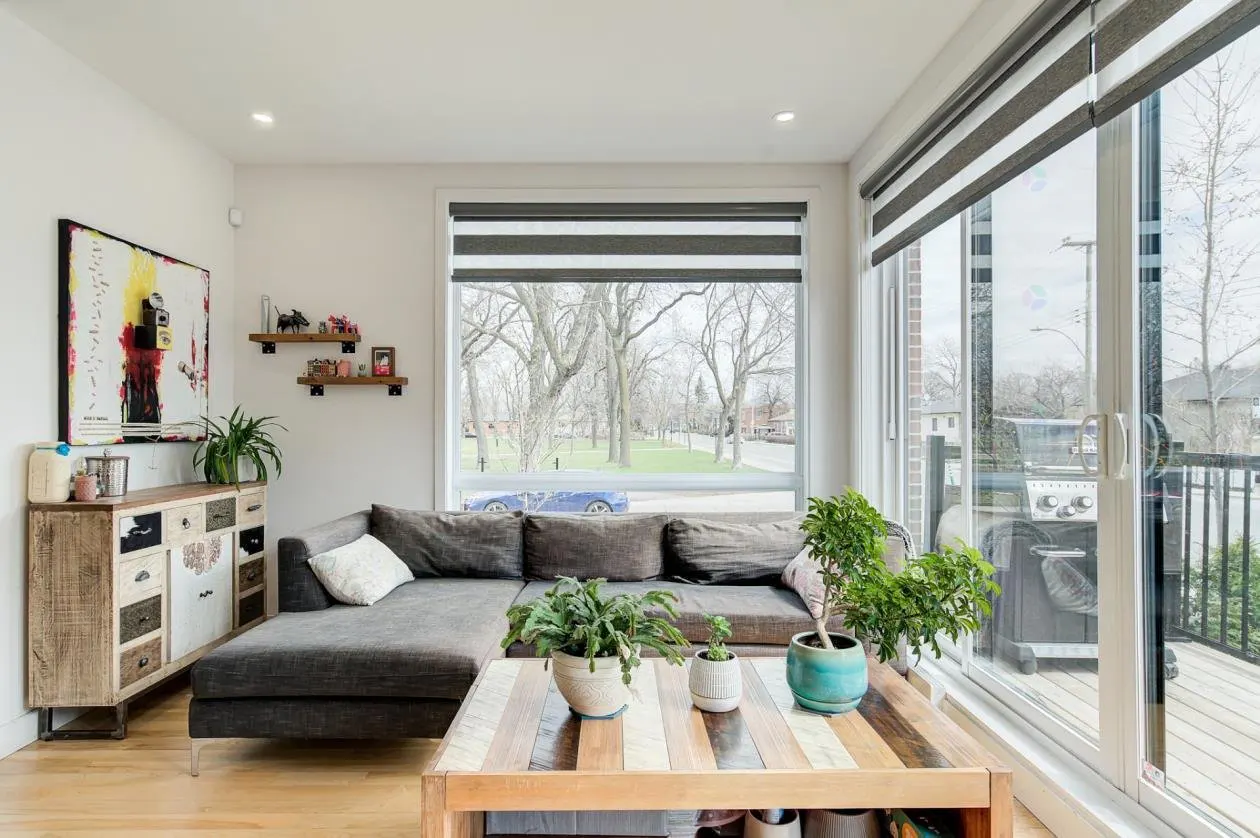 Living room with gray sectional sofa, wood coffee table with plants, and large windows with blinds. Artwork and shelves on the wall.