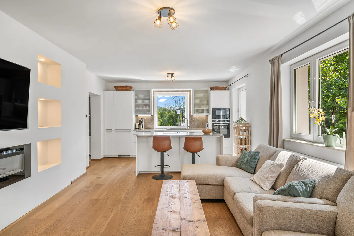 Bright, open-concept living space with a beige sectional sofa, wooden coffee table, and kitchen island with two brown stools. Large windows offer a view of greenery.