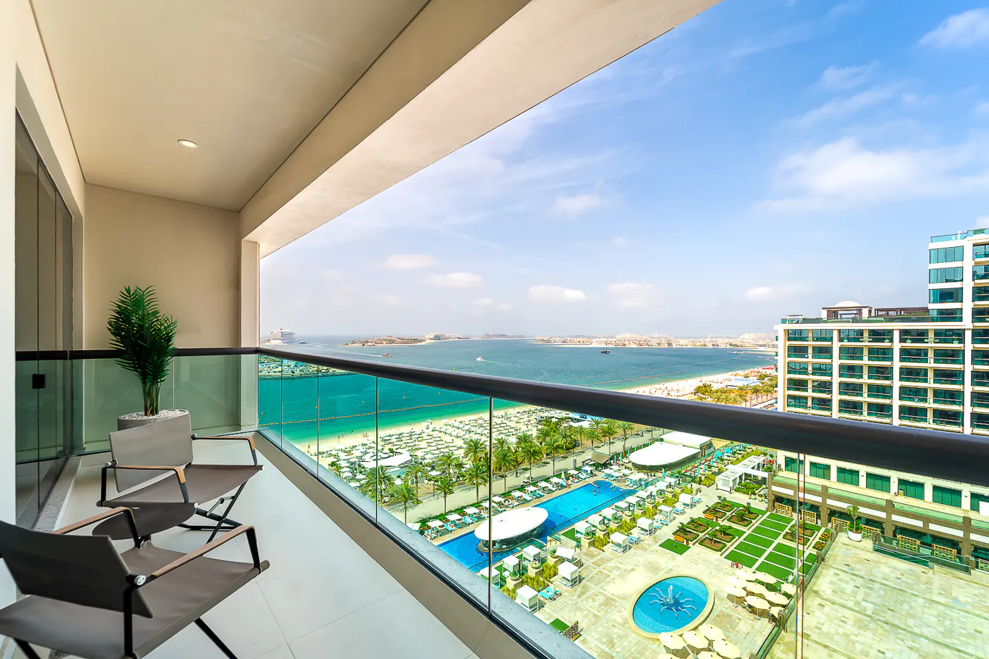 Balcony view with two chairs, a potted plant, and a glass railing overlooking a beach, pool, and ocean on a sunny day.