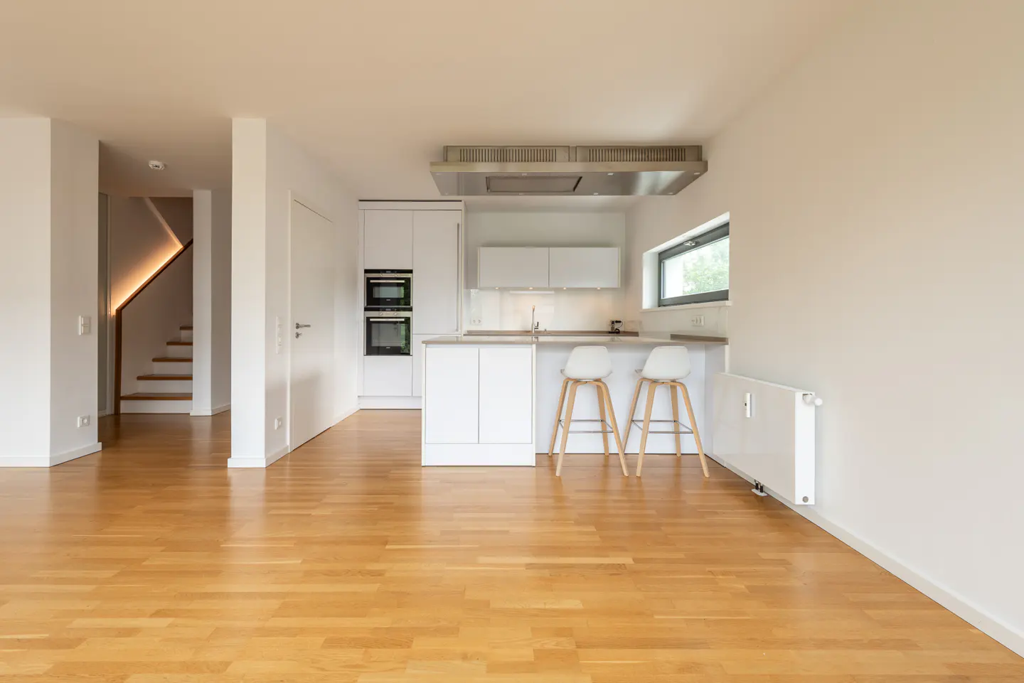 Modern white kitchen with island, two bar stools, and light wood flooring, featuring a staircase in the background.