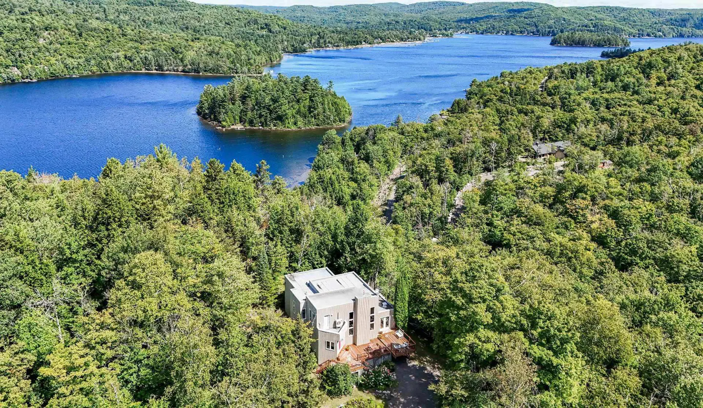 Aerial view of a modern beige house nestled in lush green trees, overlooking a blue lake with a small island.