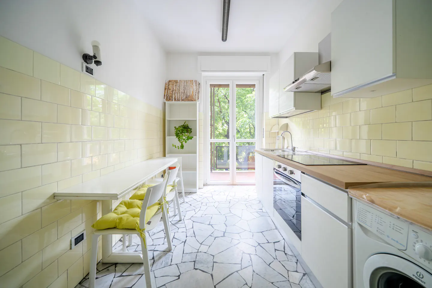 Bright, narrow kitchen with white cabinets, yellow tile backsplash, and a table with yellow cushions. A balcony door lets in natural light.