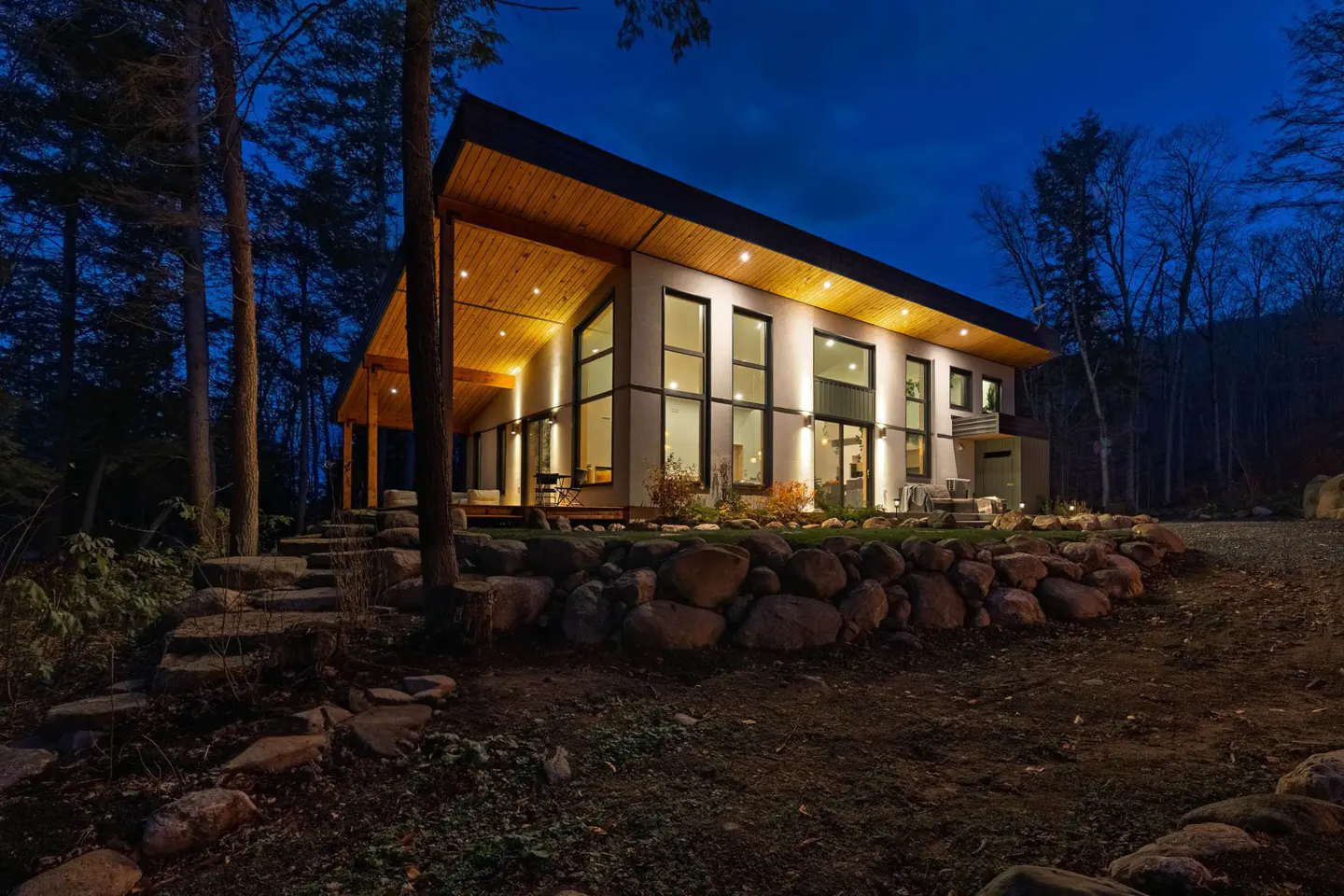 Modern home exterior at dusk. The house has a slanted roof, large windows, and a stone retaining wall. Trees surround the property.