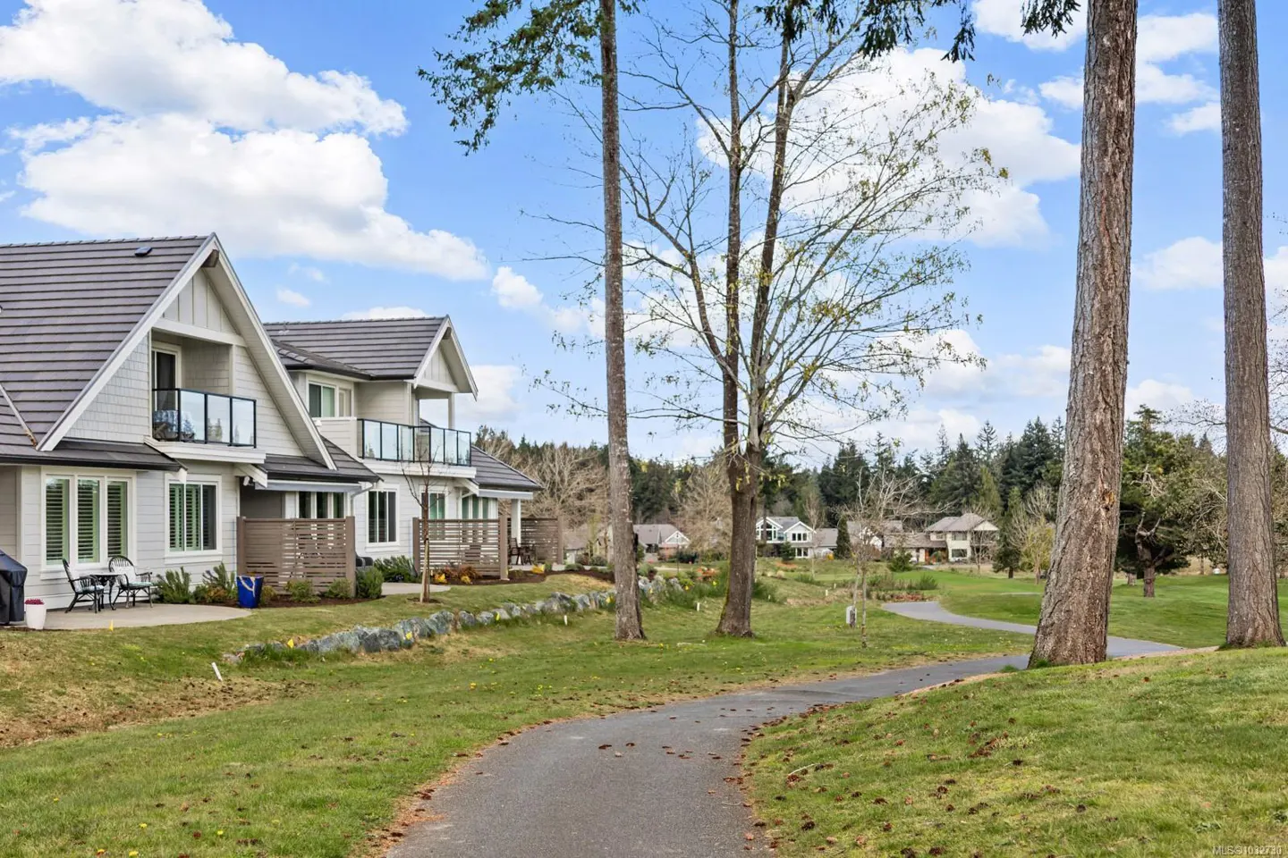 Exterior view of modern townhouses with balconies, gray roofs, and a paved path through a green lawn with trees under a blue sky.