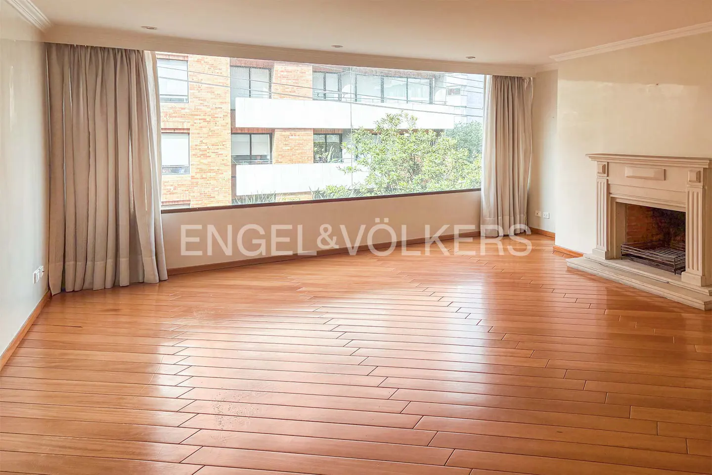 Empty living room with hardwood floors, a fireplace, and a large window with curtains. Engel & Völkers logo is visible.