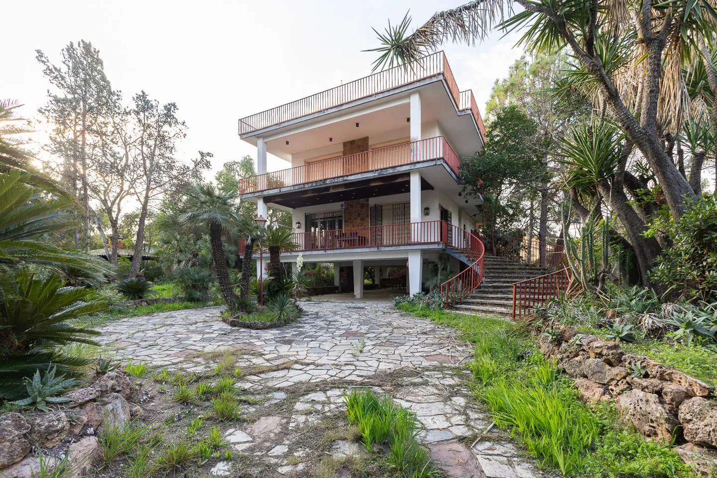 Exterior of a three-story white house with red railings, surrounded by lush greenery and a stone driveway.