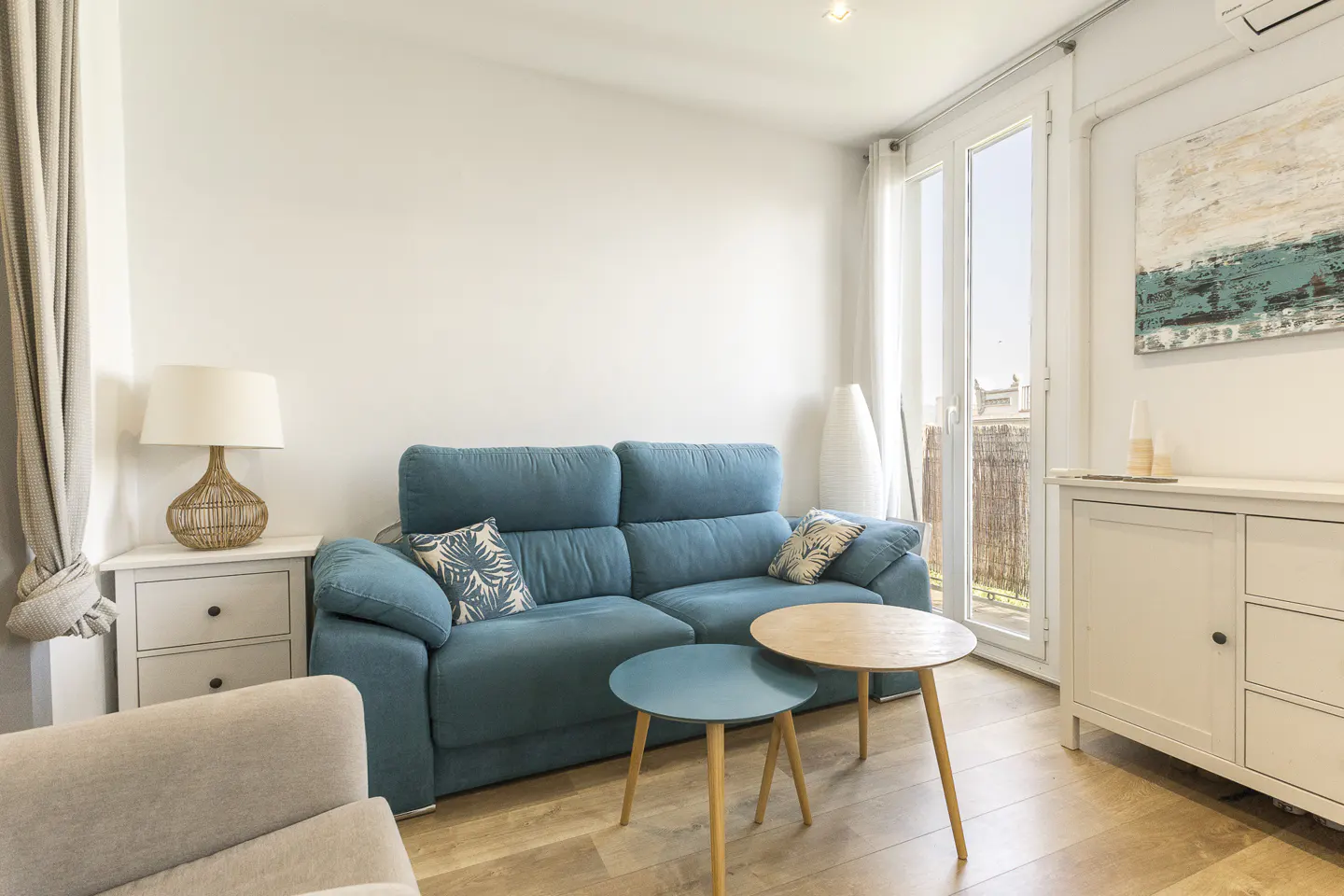 Bright living room with a blue sofa, round tables, and a white cabinet with an abstract painting above. Balcony doors let in natural light.