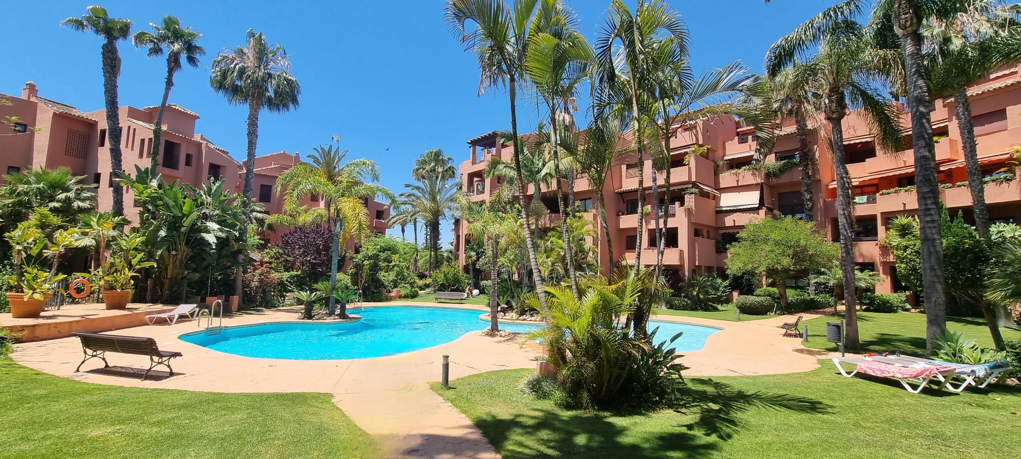 A swimming pool surrounded by lush palm trees, green lawns, and terracotta-colored apartment buildings under a blue sky.