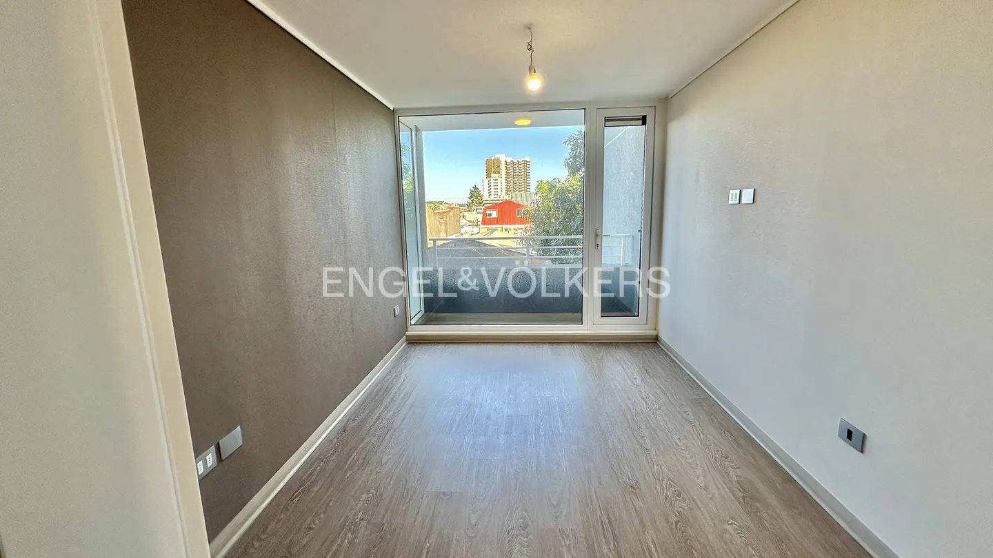 Empty room with wood floors, white walls, and a brown accent wall. Balcony with city view through sliding glass doors.