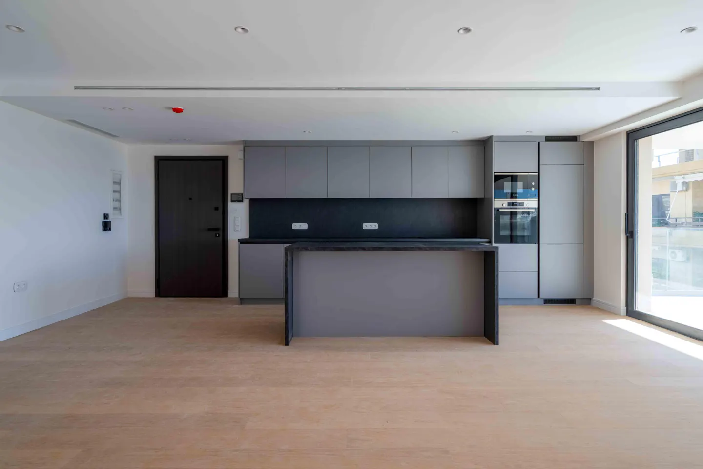 A modern kitchen with gray cabinets, a black countertop, and a kitchen island. A black door is on the left, and a sliding glass door is on the right.