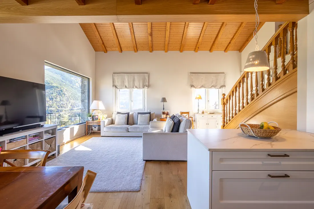 Bright living room with wood floors, white sofas, and a large window with a mountain view. A wooden staircase is visible.