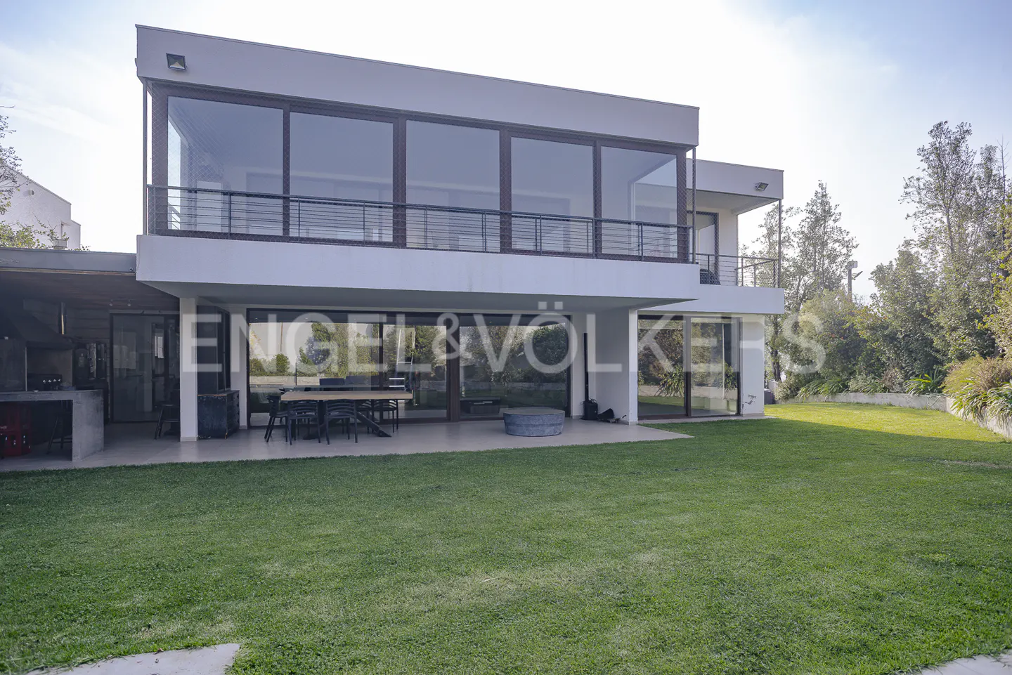 Two-story modern house with large windows, balcony, patio, and green lawn. Trees in the background.