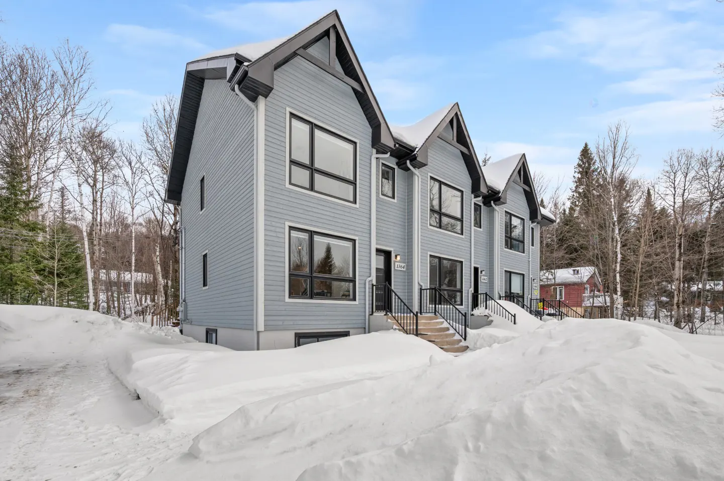 Three light blue townhouses with black trim and large windows sit on a snow-covered lot under a blue sky.