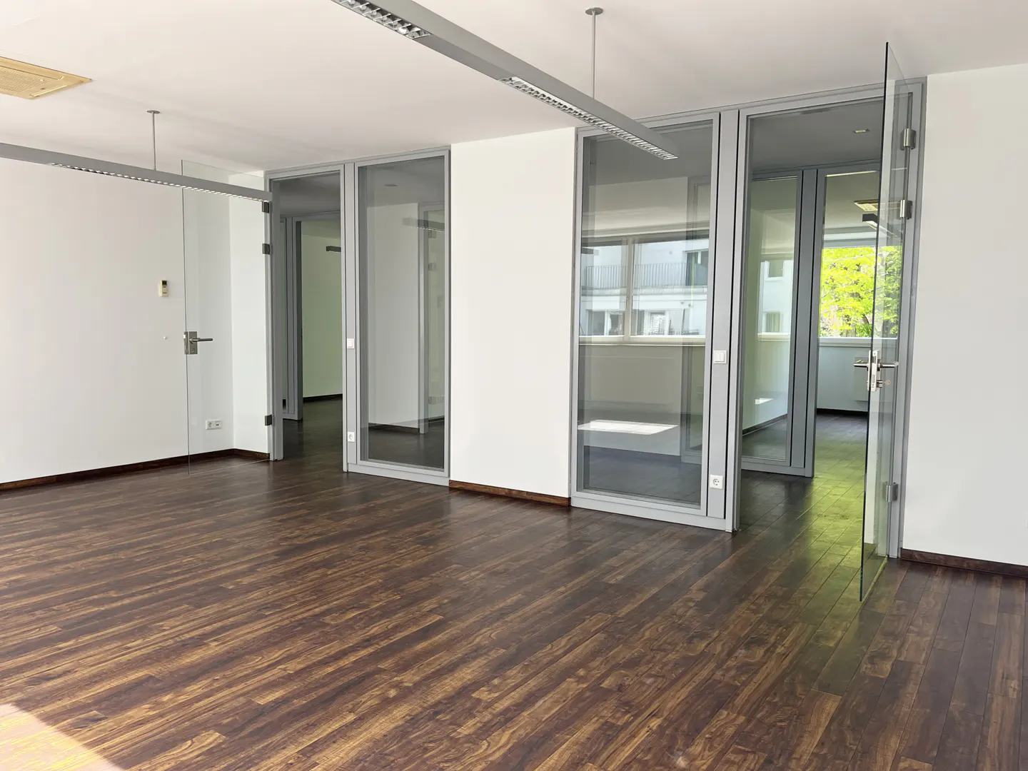 Empty office space with dark wood floors, white walls, and glass-walled rooms. Overhead lighting is visible.