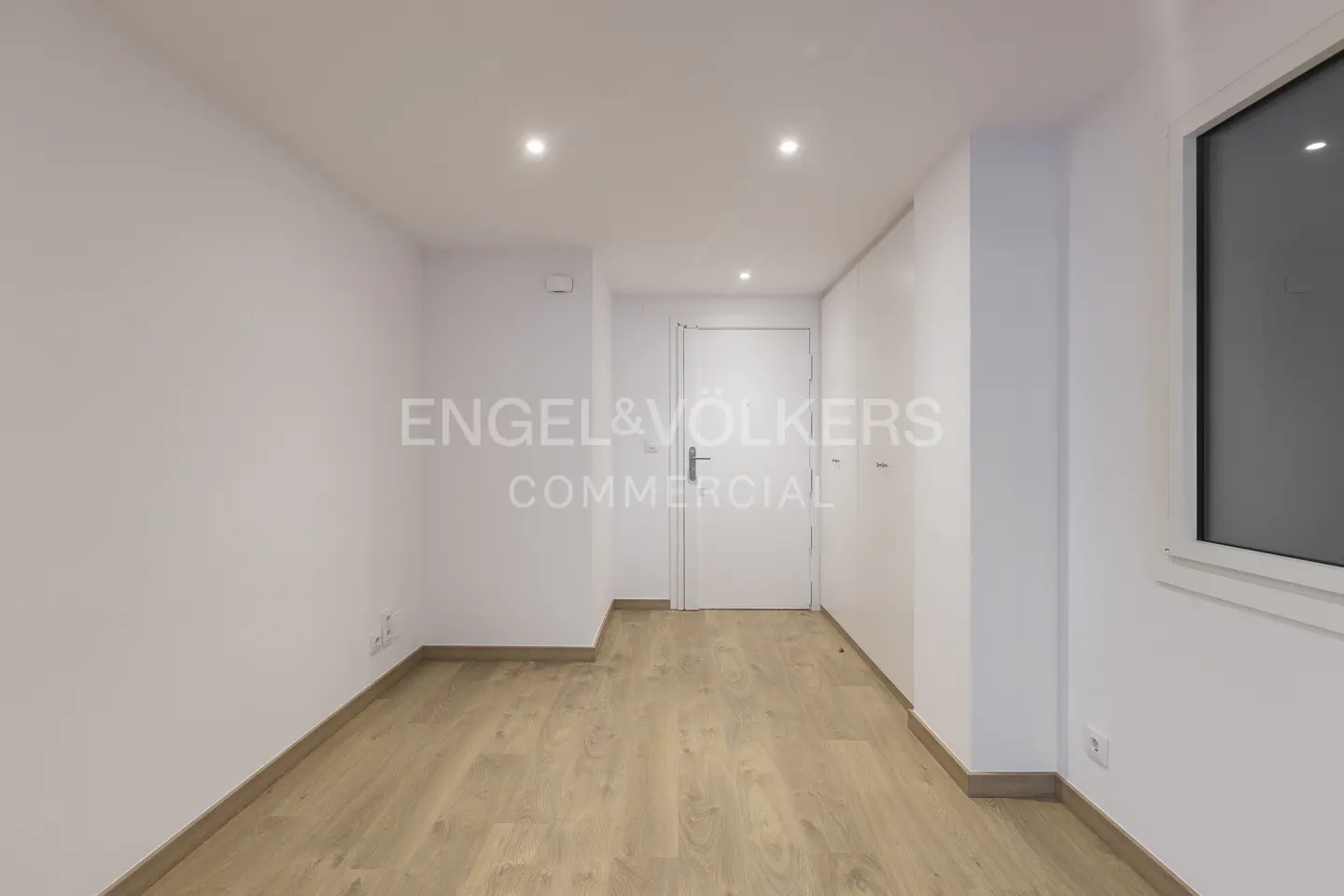 Empty room with white walls, light wood flooring, recessed ceiling lights, a white door, and a window on the right.