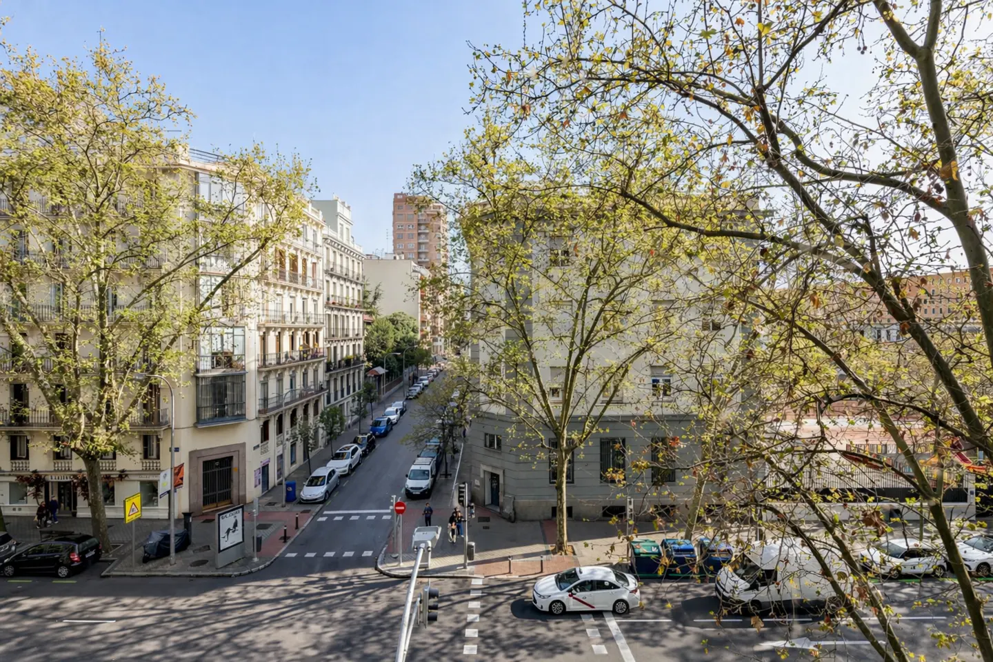 Street view of Madrid, Spain, with beige buildings, trees, and cars on a sunny day.