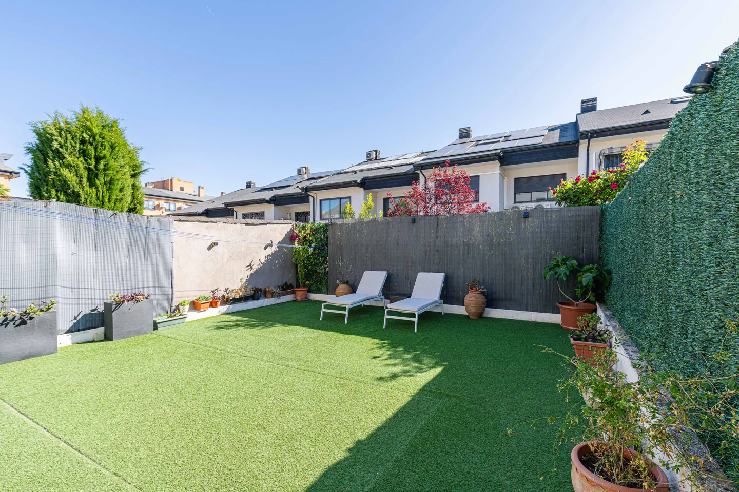 A backyard with artificial grass, two lounge chairs, and a fence with plants. Row houses are in the background.